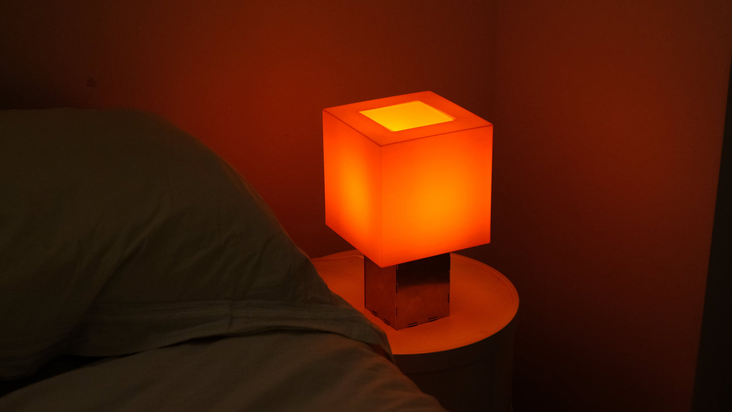 Square lamp with a warm glow on a bedside table in a dimly lit room.